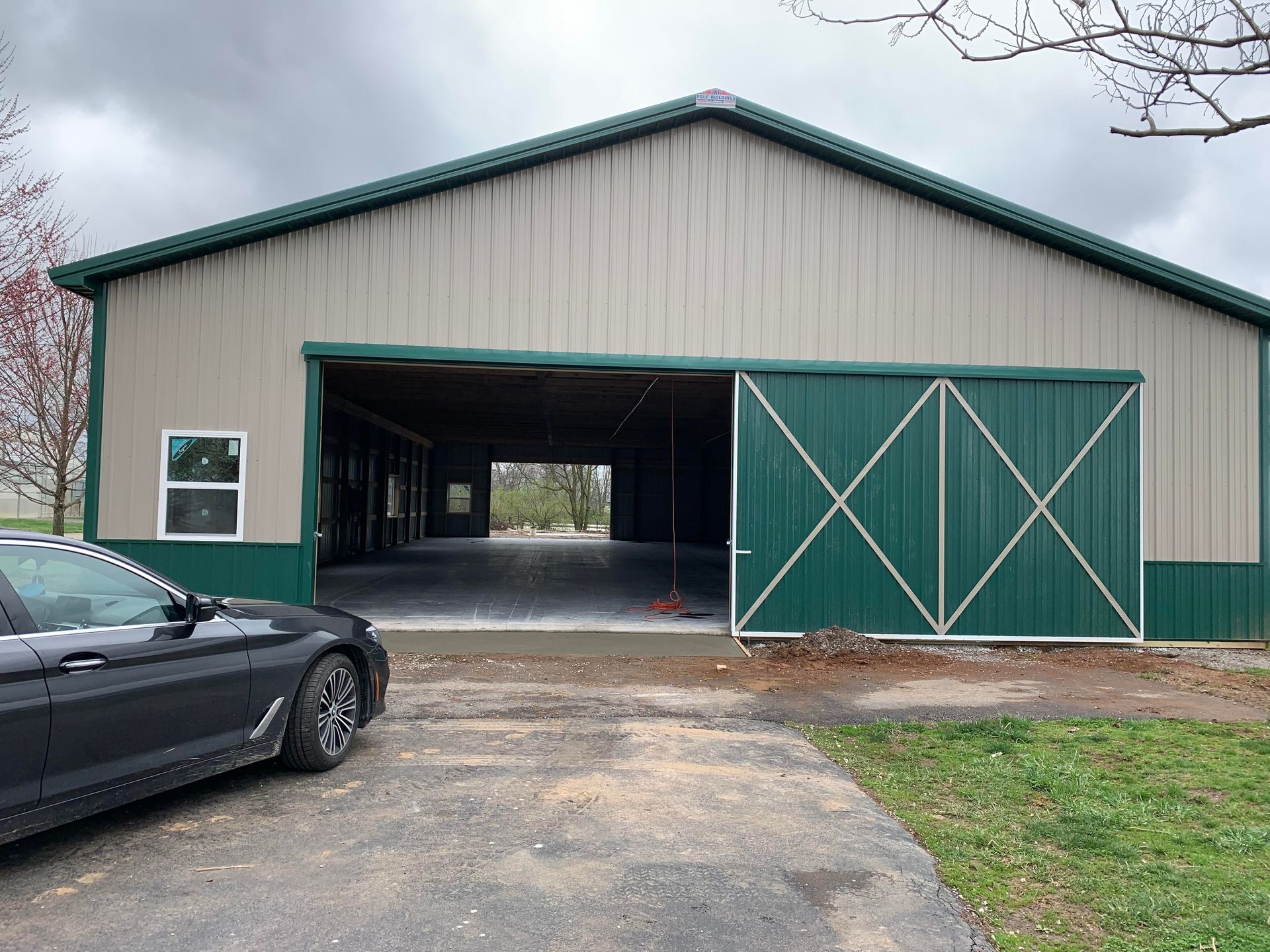 A large metal-sided building with a wide, open bay door and a green sliding barn door, beside a parked dark sedan.