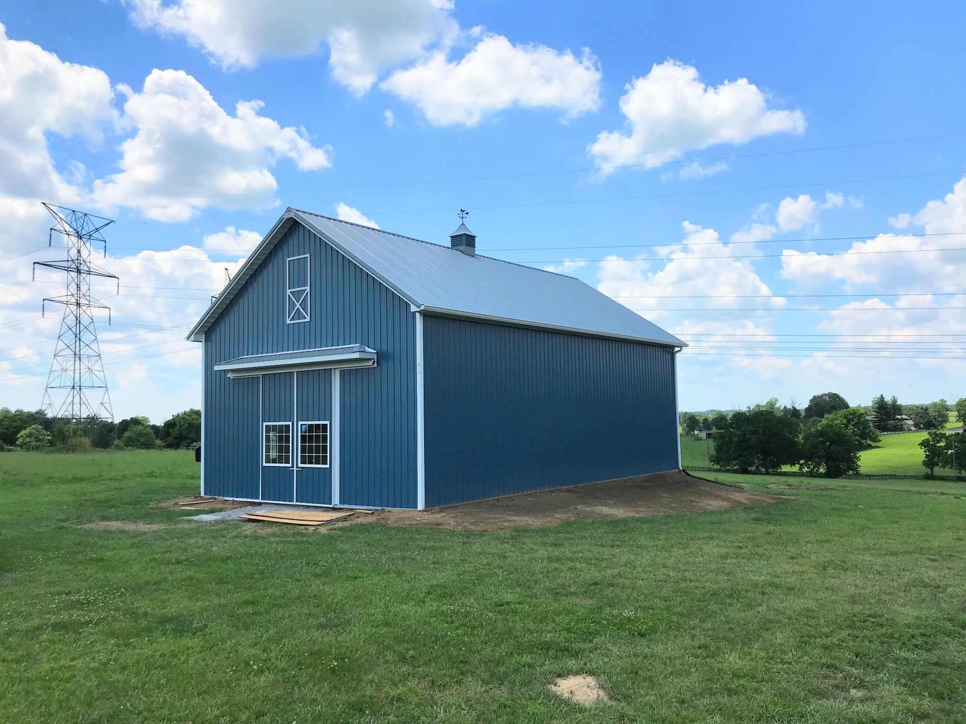 A blue metal barn with white trim and a silver roof sits in a green field under a blue sky with fluffy white clouds.