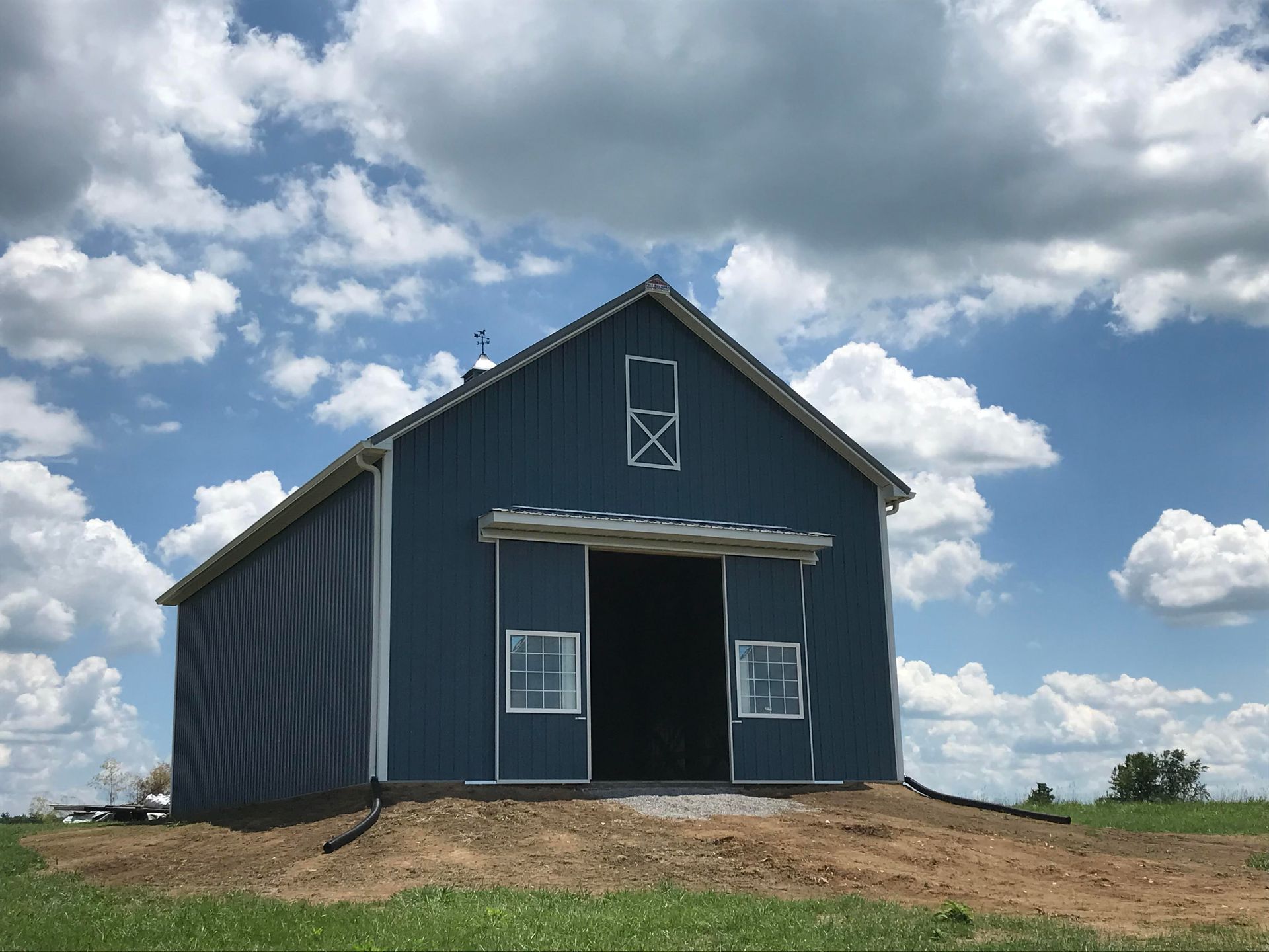 A blue barn with a large open doorway, white trim, and a small window above, set in a field under a cloudy blue sky.