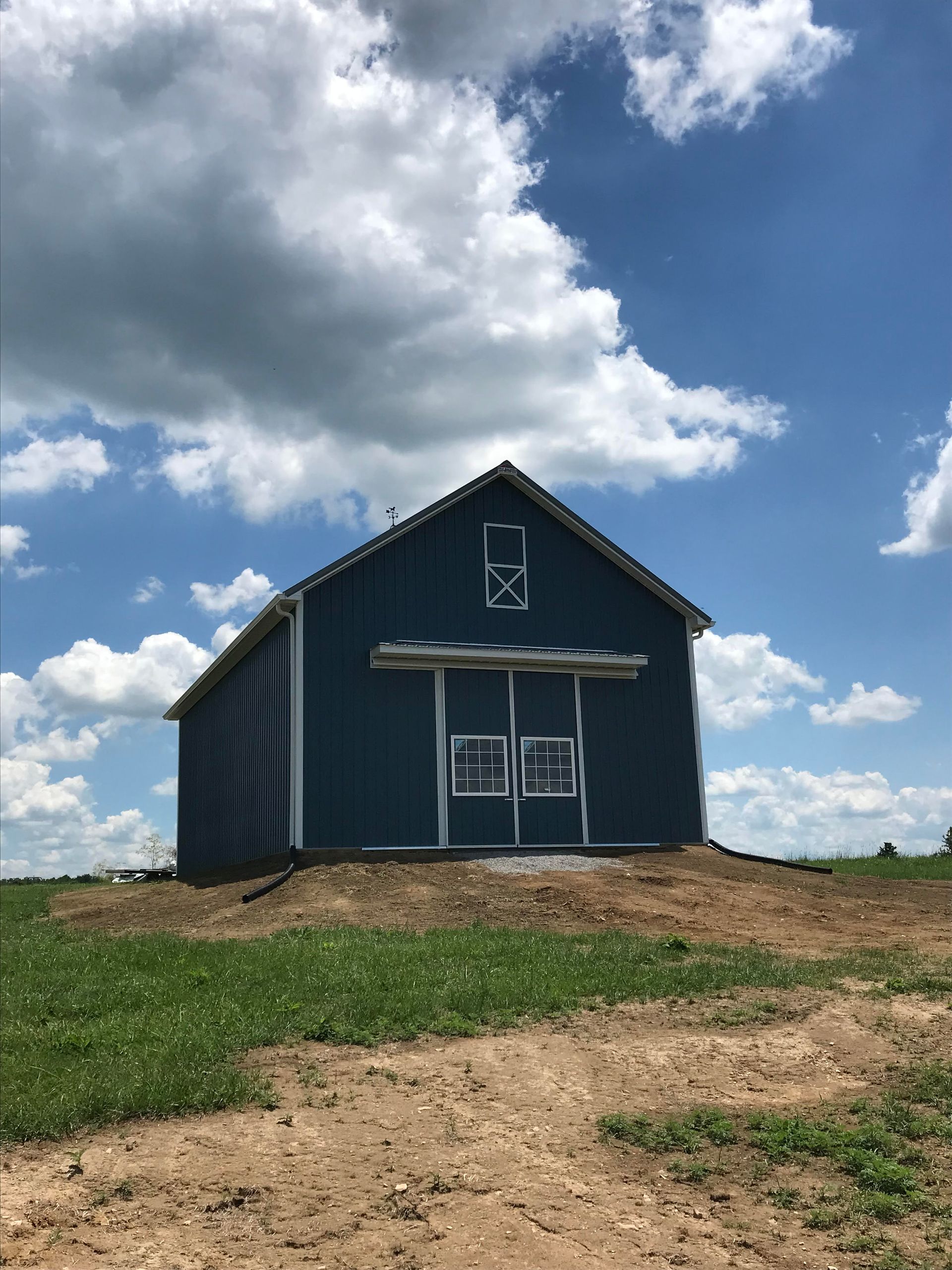 A blue barn stands on a dirt mound in a grassy field under a bright blue sky with large white clouds.
