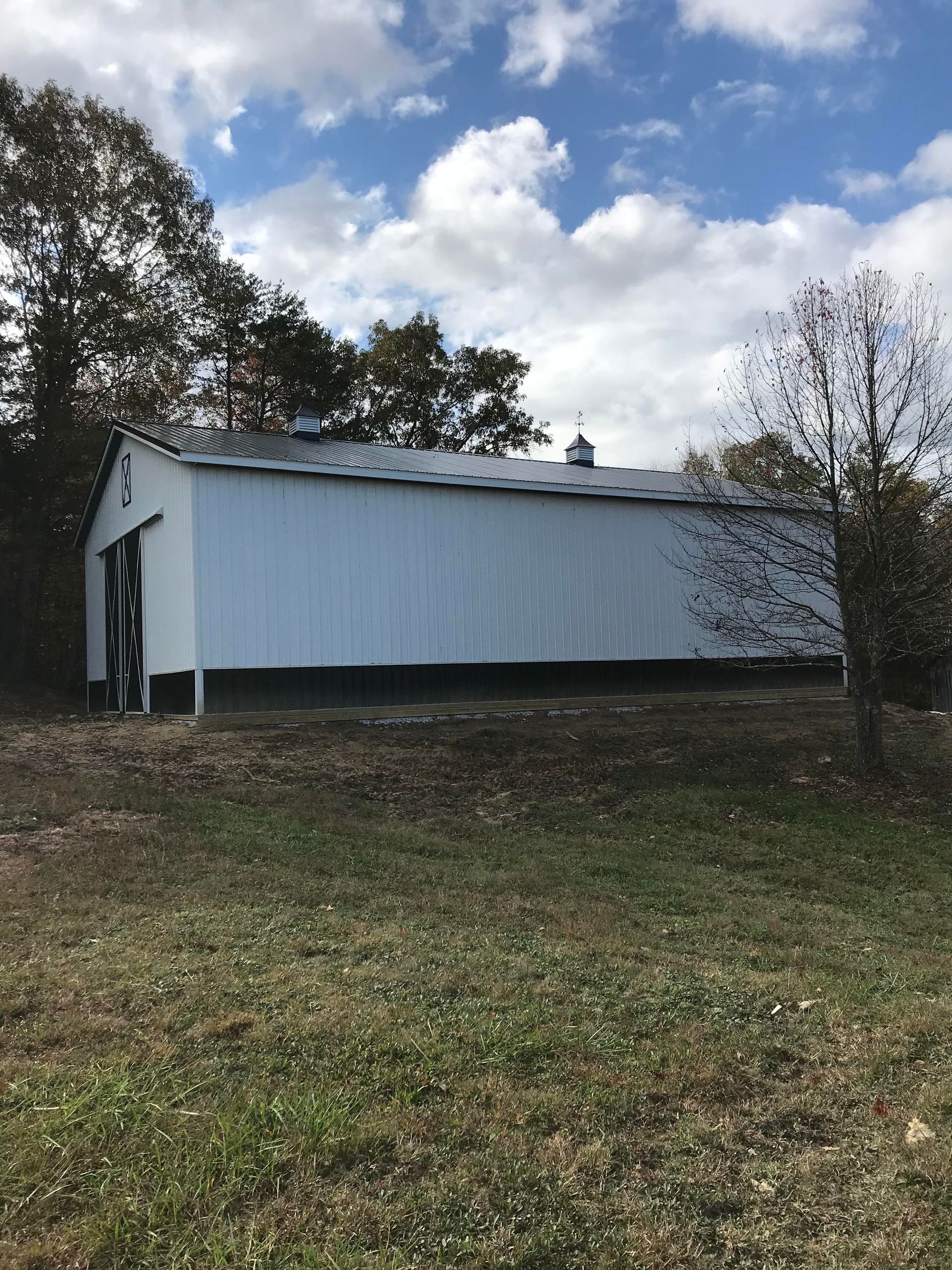 A long white pole barn with a dark trim stands in a grassy field under a bright, cloudy sky.