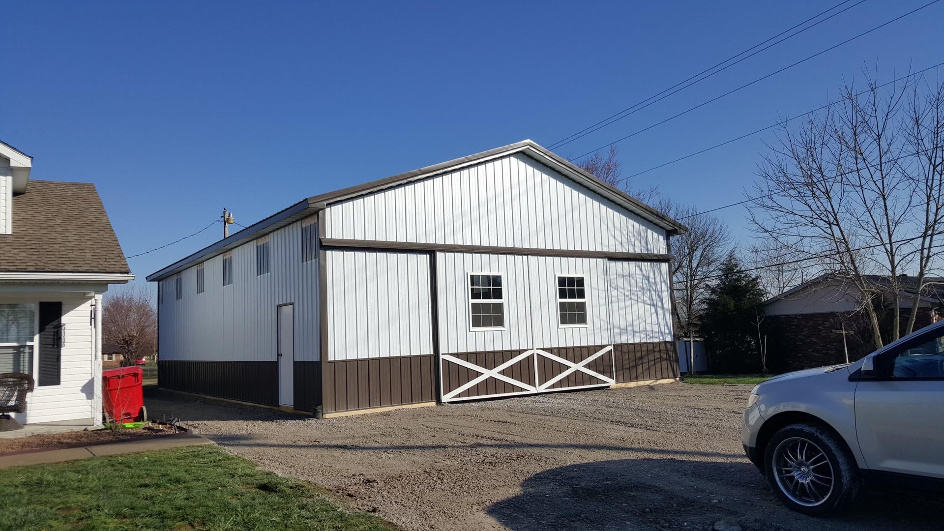 A white, metal-sided barn with brown trim and X-bracing on the sliding doors sits next to a house on a gravel lot.