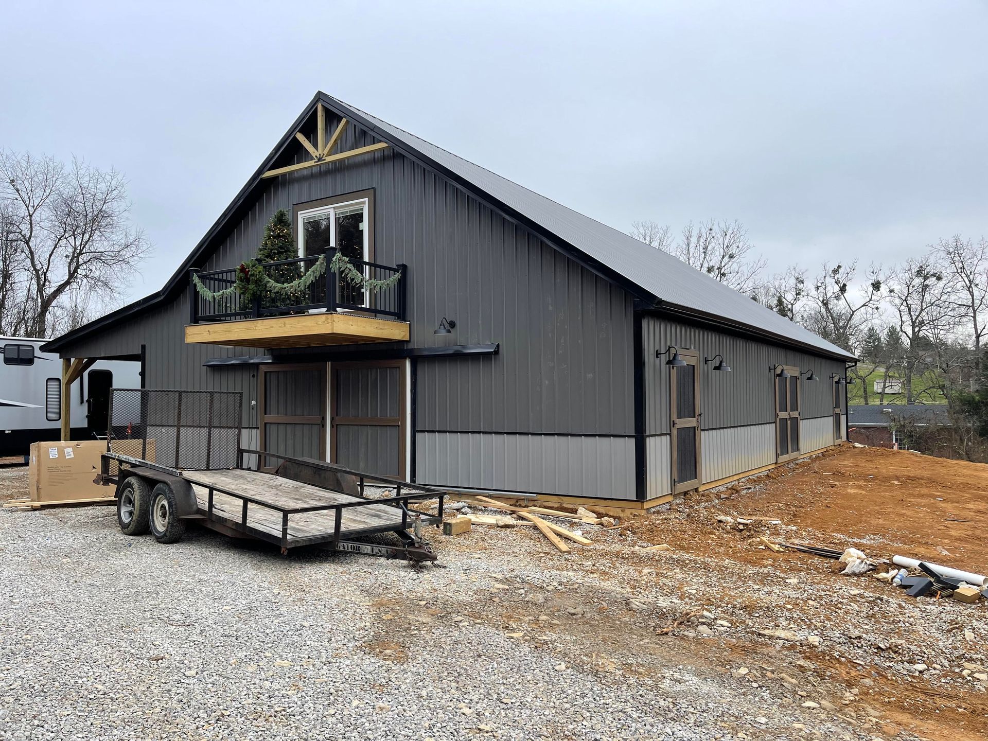 A large, dark grey metal barn building with a balcony and sliding doors, situated on a gravel lot with a utility trailer.