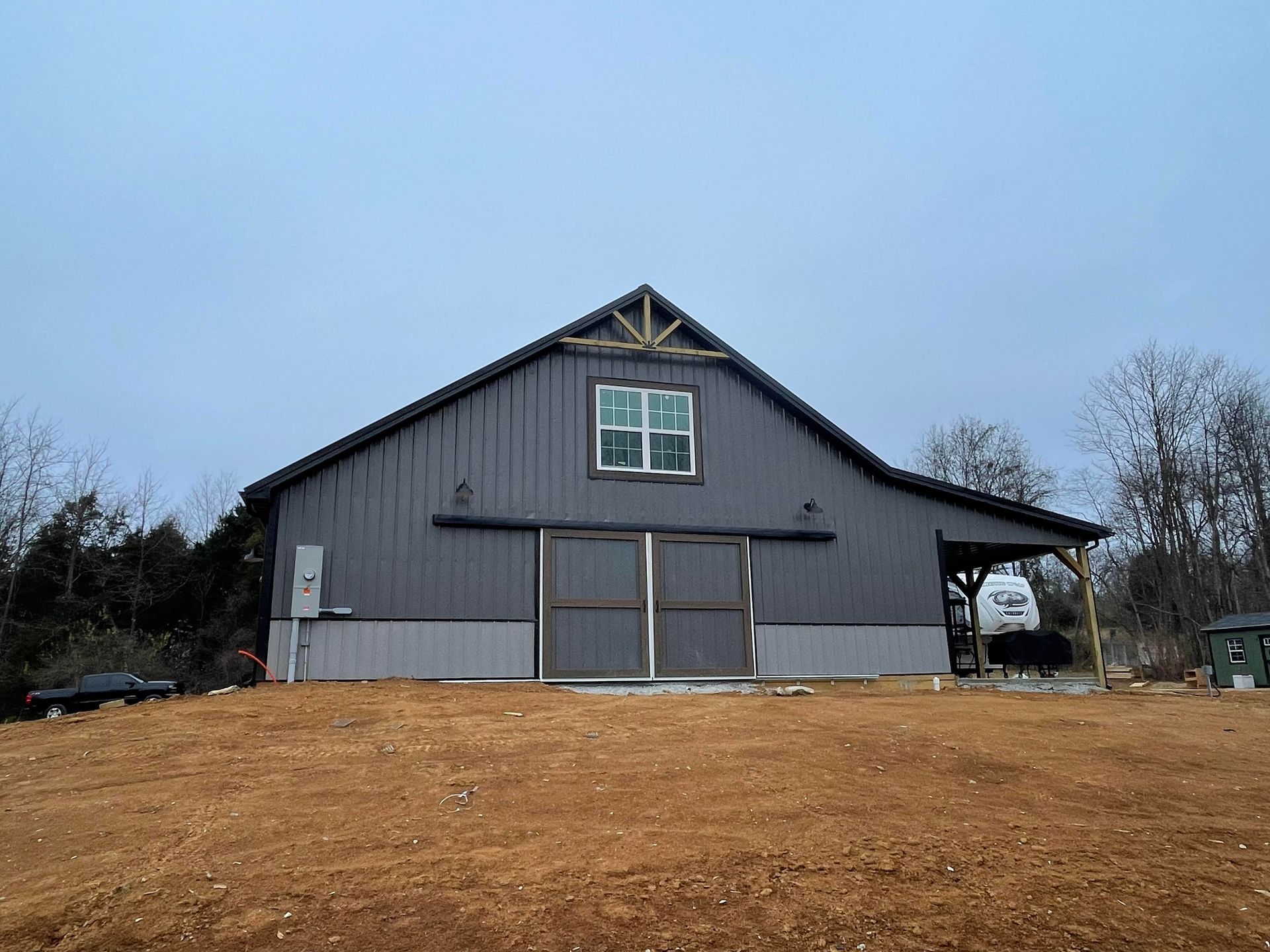 A gray metal barn with sliding double doors, a loft window, and a side lean-to, situated on a dirt lot under a pale sky.