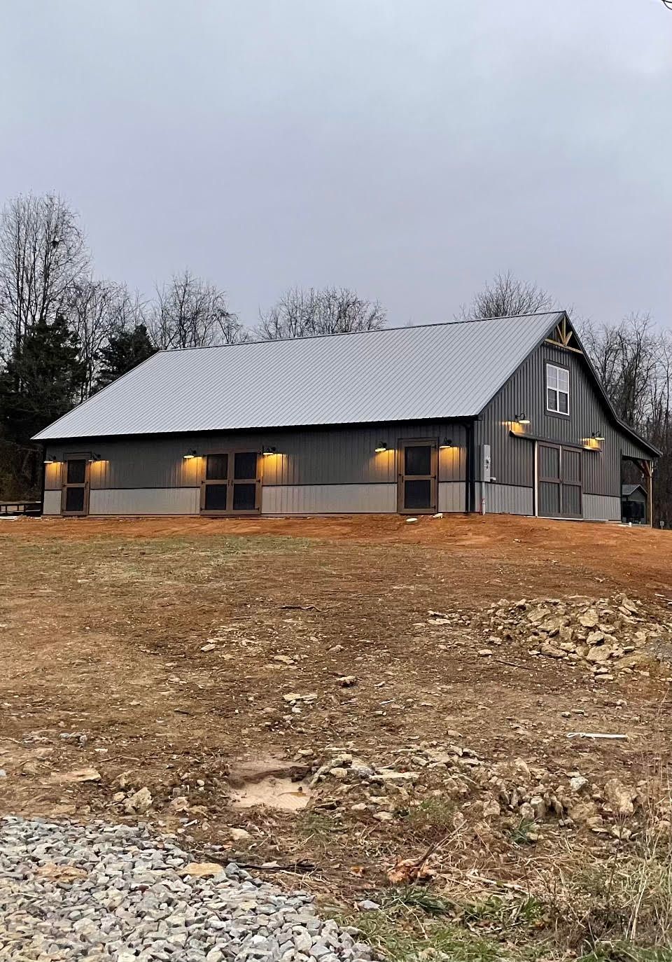 A gray metal barn with an angled roof and exterior lighting, situated on a grassy hill under a cloudy sky.