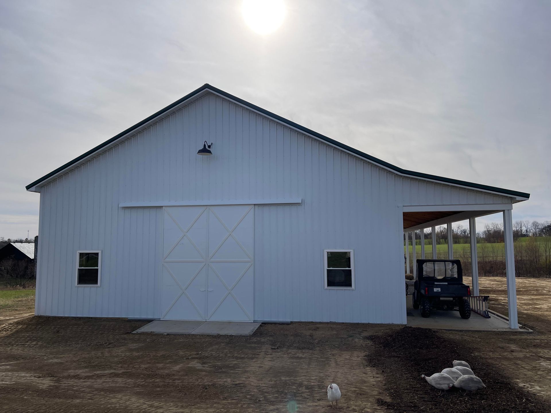 A white pole barn with a dark roof, sliding center doors, two windows, and an open side porch housing an ATV.