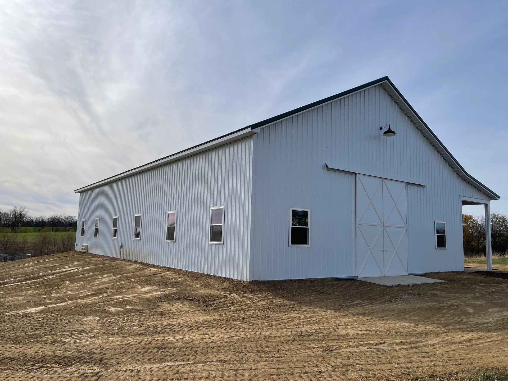 A large, white metal pole barn with several windows and sliding doors, set on a dirt lot under a blue sky.