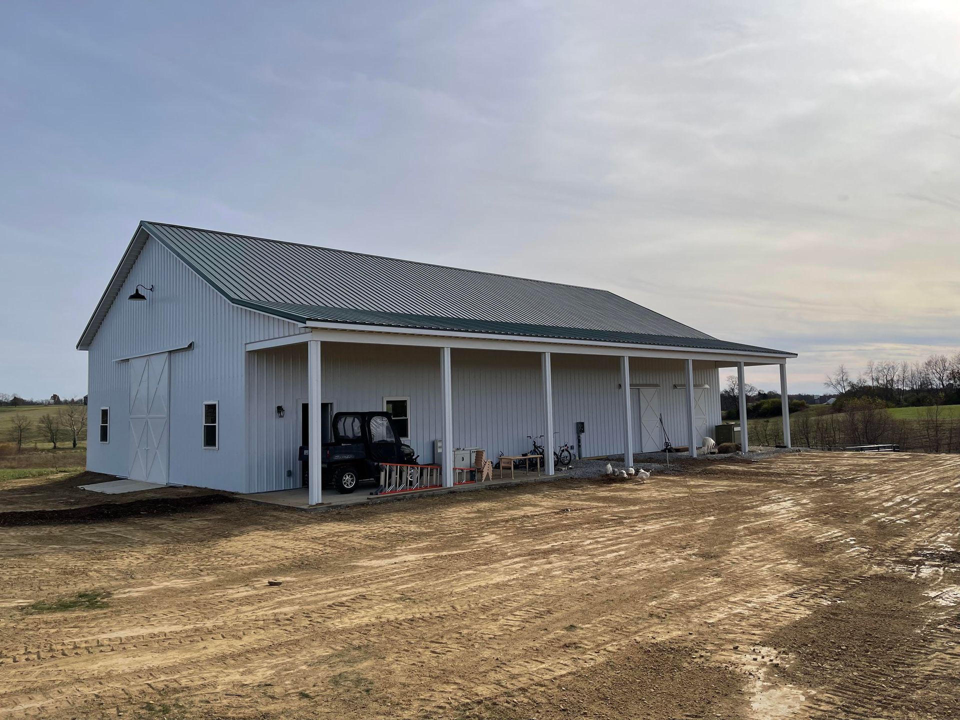 A white, board-and-batten pole barn with a dark metal roof and an open porch sits on a dirt lot under a sunny sky.