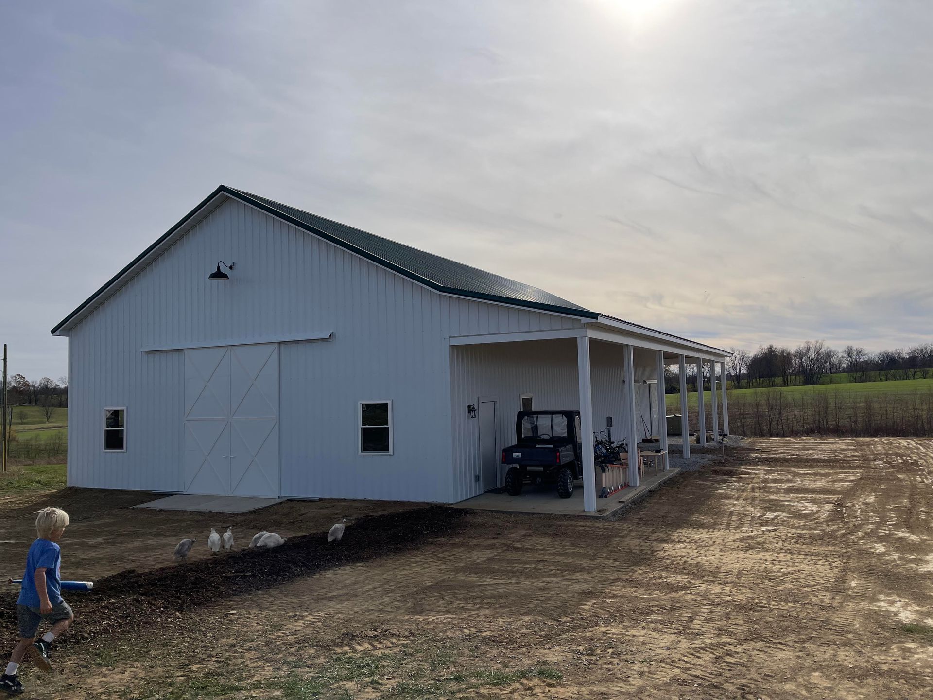 A white barn with a green metal roof and an open-sided porch, next to an all-terrain vehicle on a dirt lot.