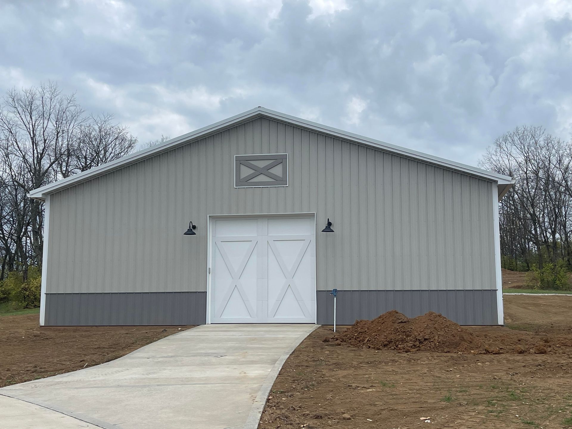 A light gray pole barn with a dark gray wainscot, white double doors, and a small vent on a concrete driveway.