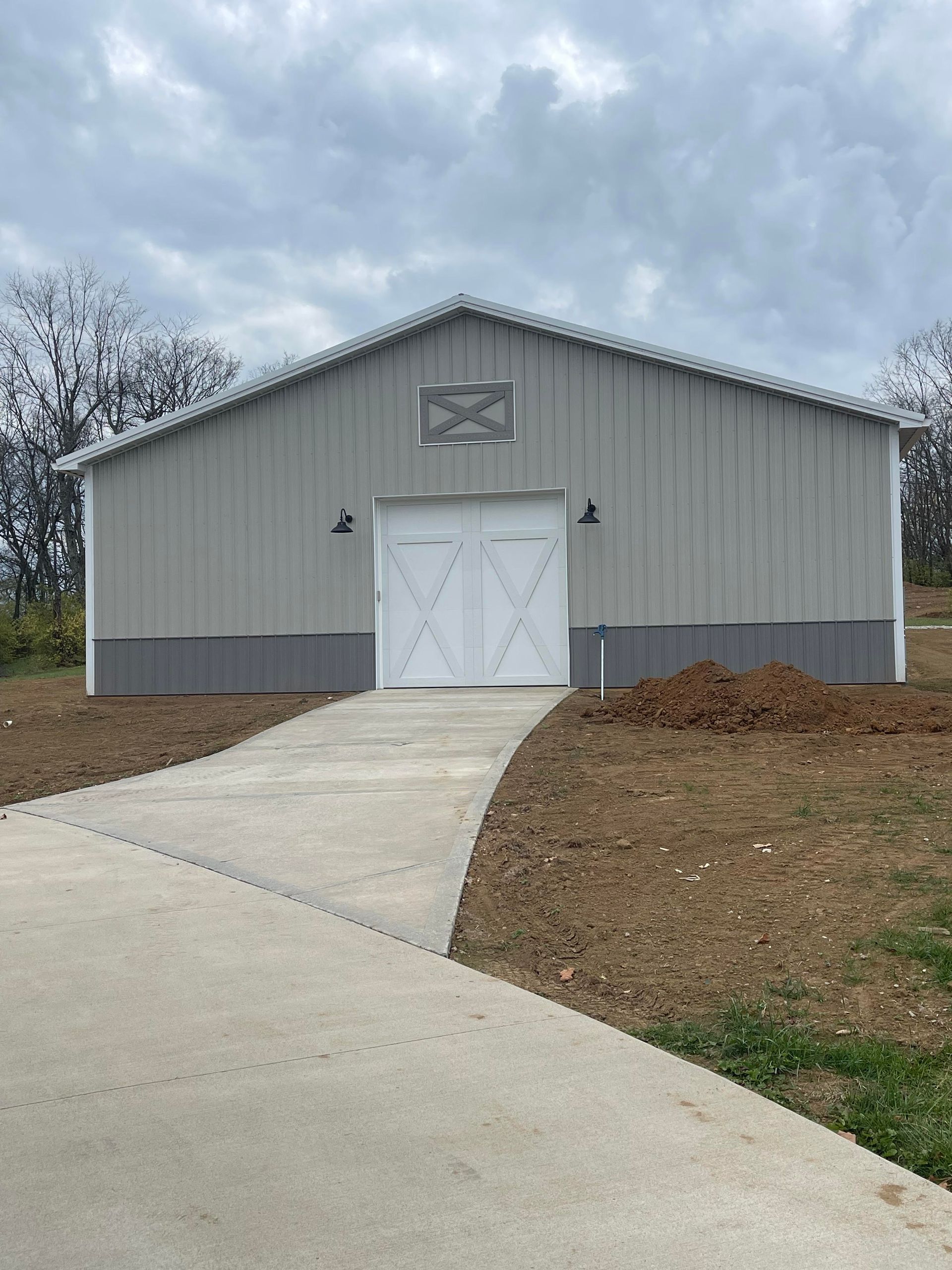A light gray metal barn with white double doors, a small window above, and a concrete driveway leading to the entrance.