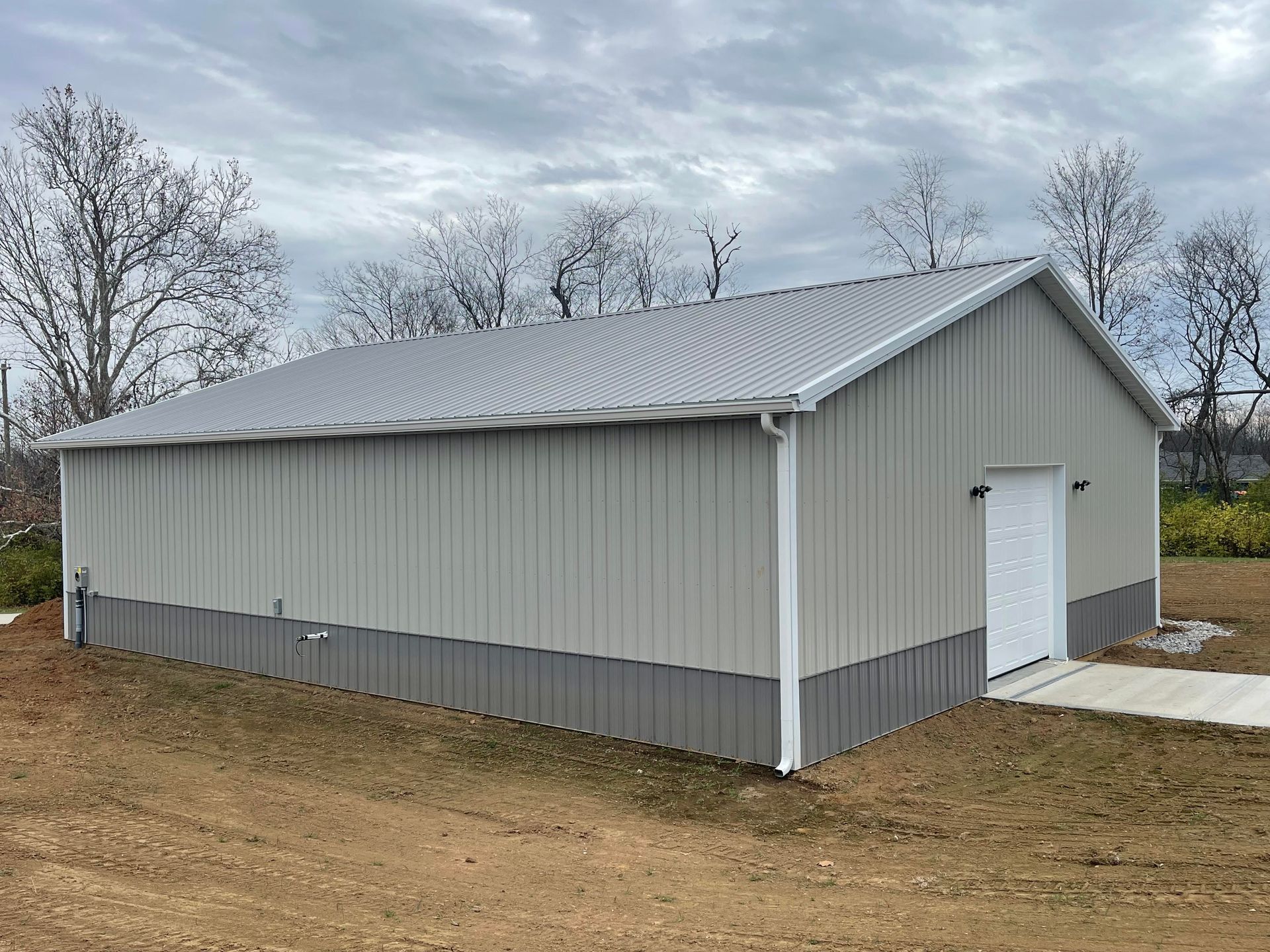 A light gray metal building with a dark gray base, white trim, and a side entrance door in a grassy, rural setting.