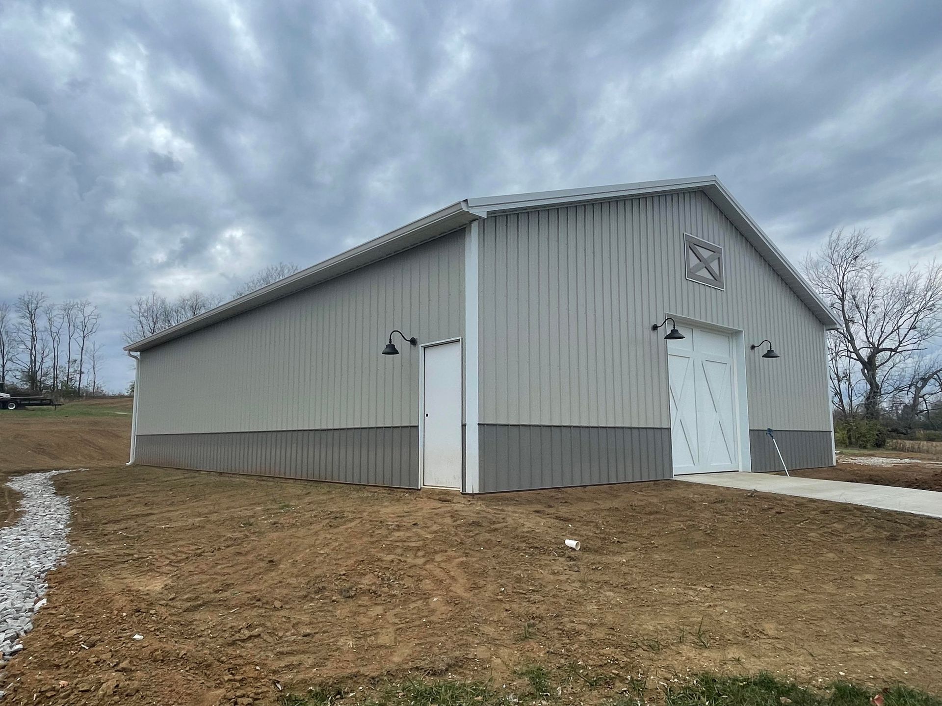 A light grey metal barn with a dark grey wainscot, white doors, and black sconce lights under a cloudy sky.