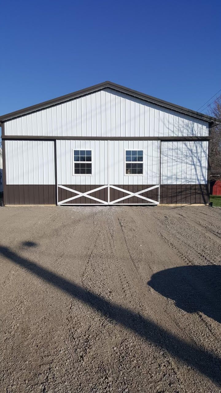 A white metal barn with dark brown trim, two windows, and X-patterned sliding doors sits on a gravel lot under blue sky.