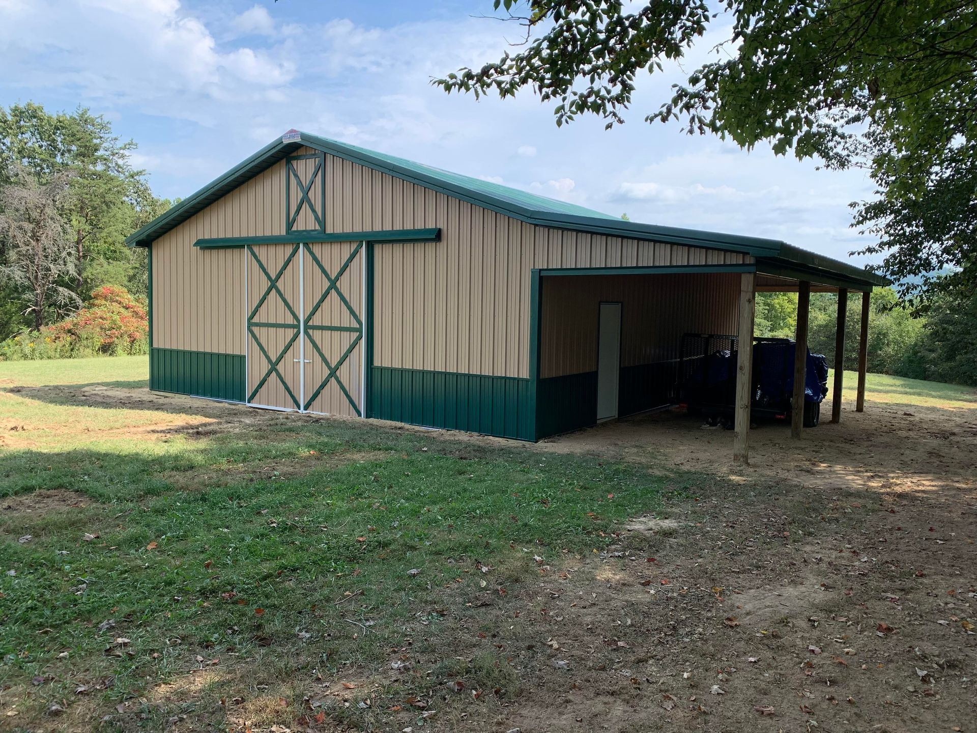 Tan and green metal barn with a sliding door and an attached side lean-to, situated in a grassy field.