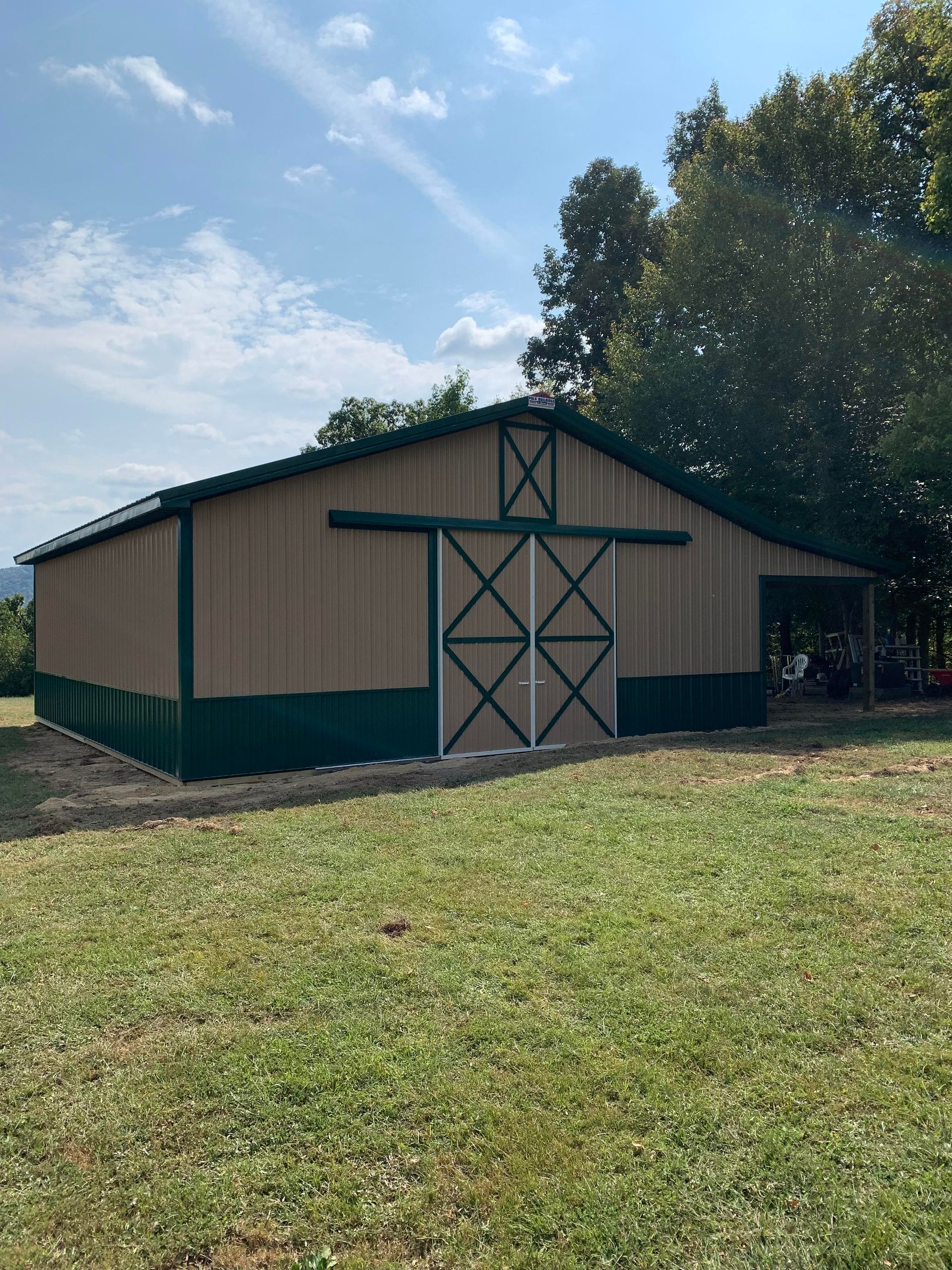 A tan and dark green barn with sliding double doors, set in a grassy field under a sunny blue sky.