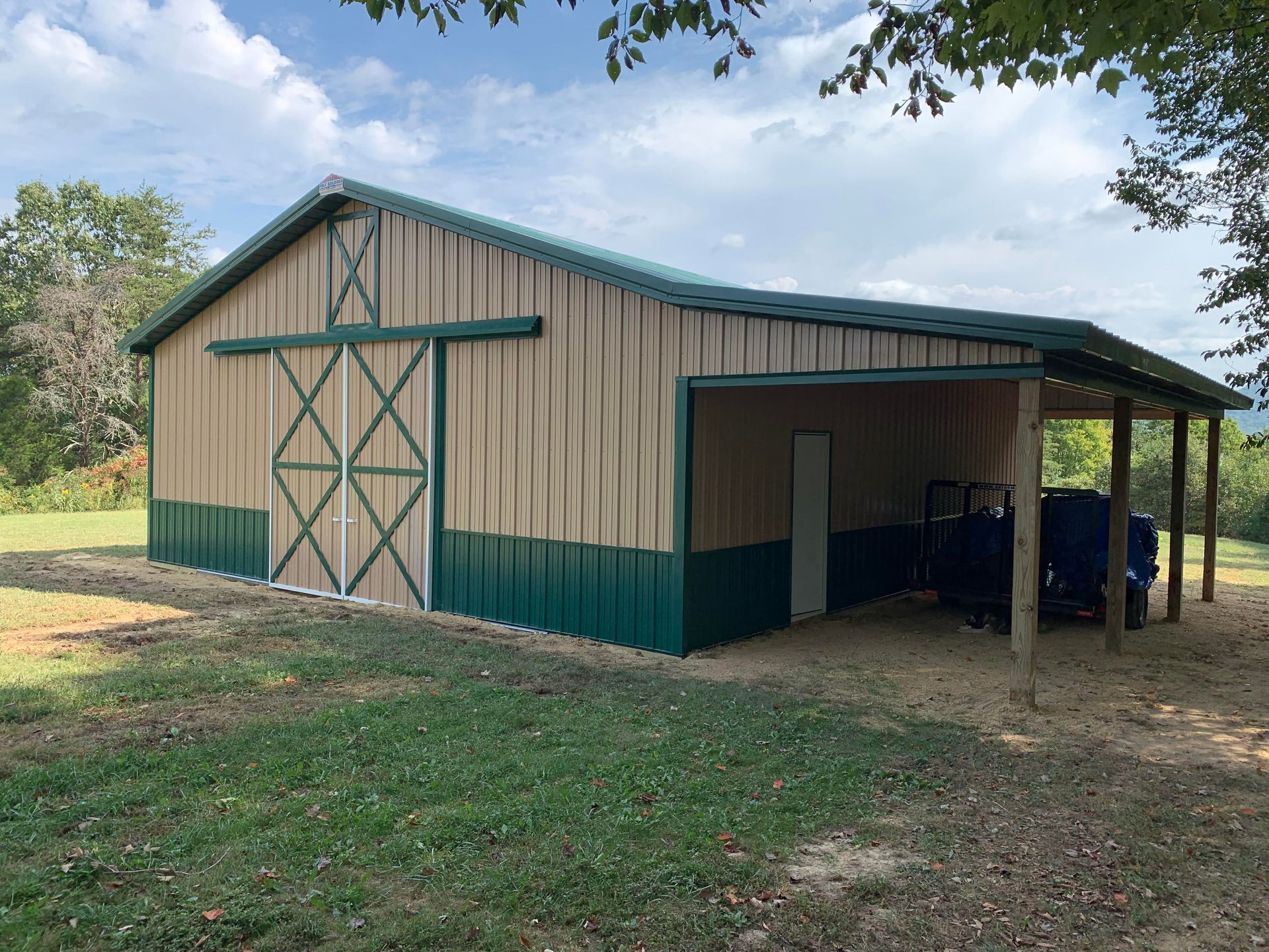 A tan and green metal barn with sliding double doors and an open-sided lean-to, situated in a grassy field.