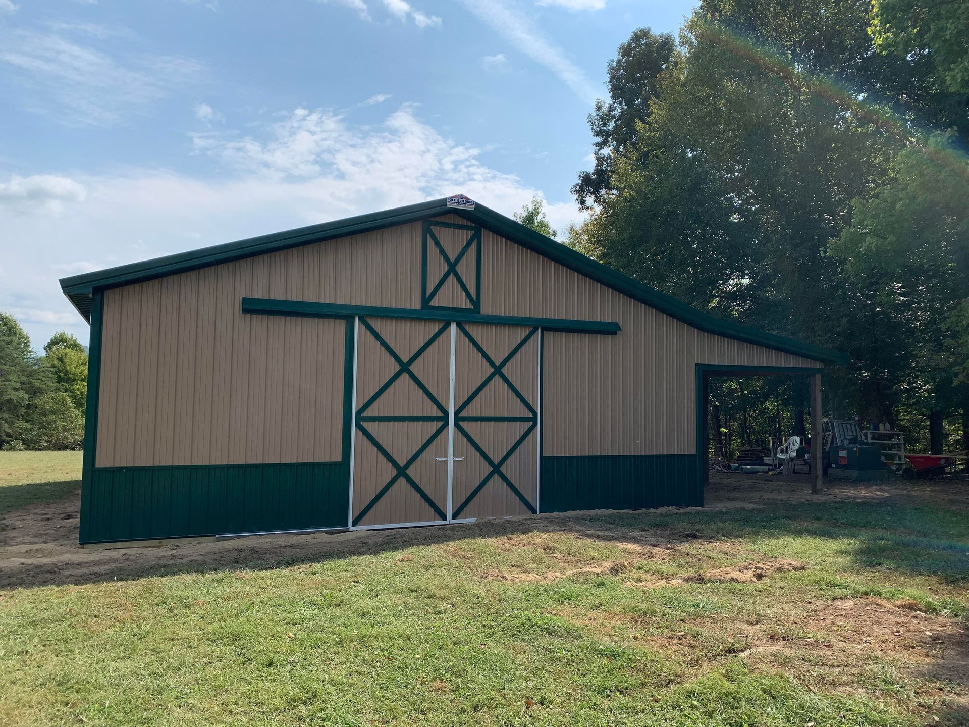 Tan and green pole barn with sliding doors and an attached side porch on a grassy lot under a blue sky.