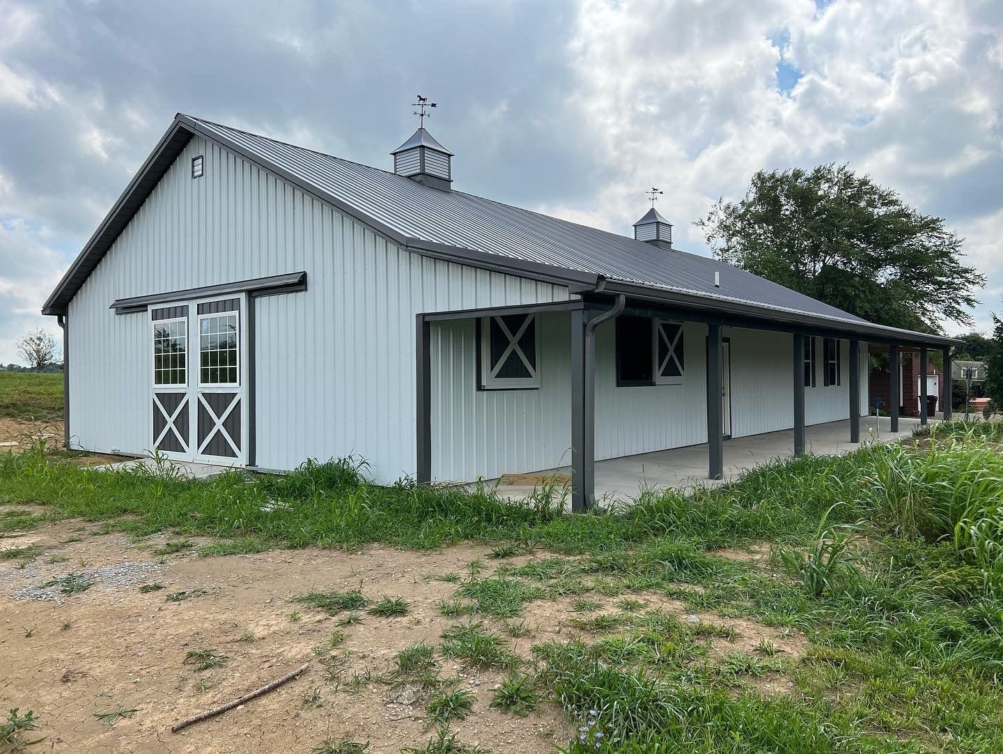A white metal barn with a grey roof, sliding doors, and a covered porch stands in a grassy field under a cloudy sky.
