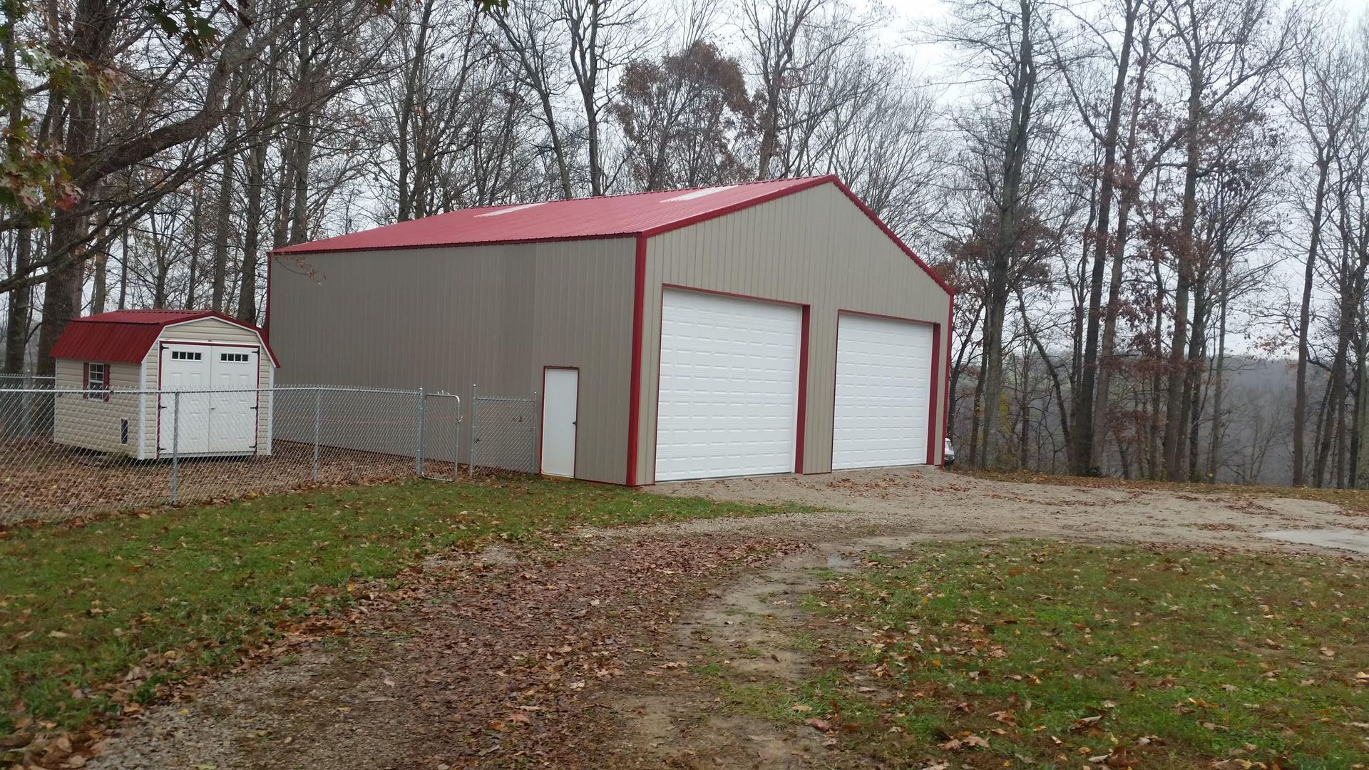A tan metal garage with two white bay doors and a red roof sits on a gravel lot next to a small shed near a forest.