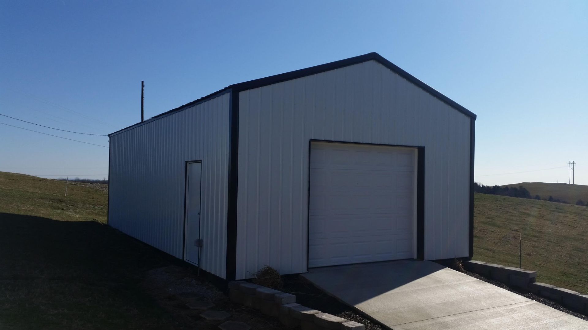 A white metal garage with a black trim and a roll-up door stands on a grassy hillside under a clear blue sky.