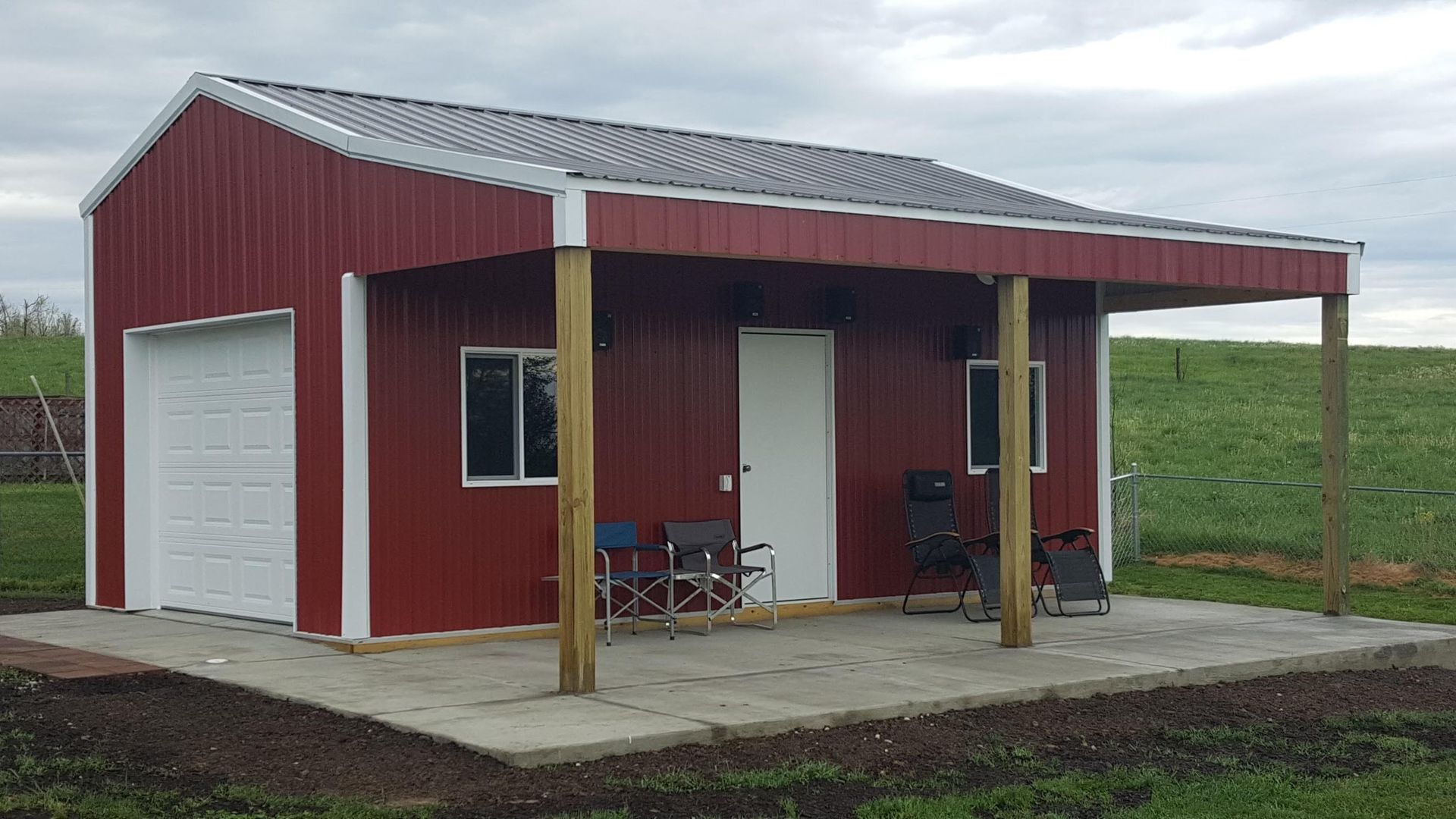 A red metal shed with a white garage door, a side door, two windows, and a covered front porch on a concrete slab.