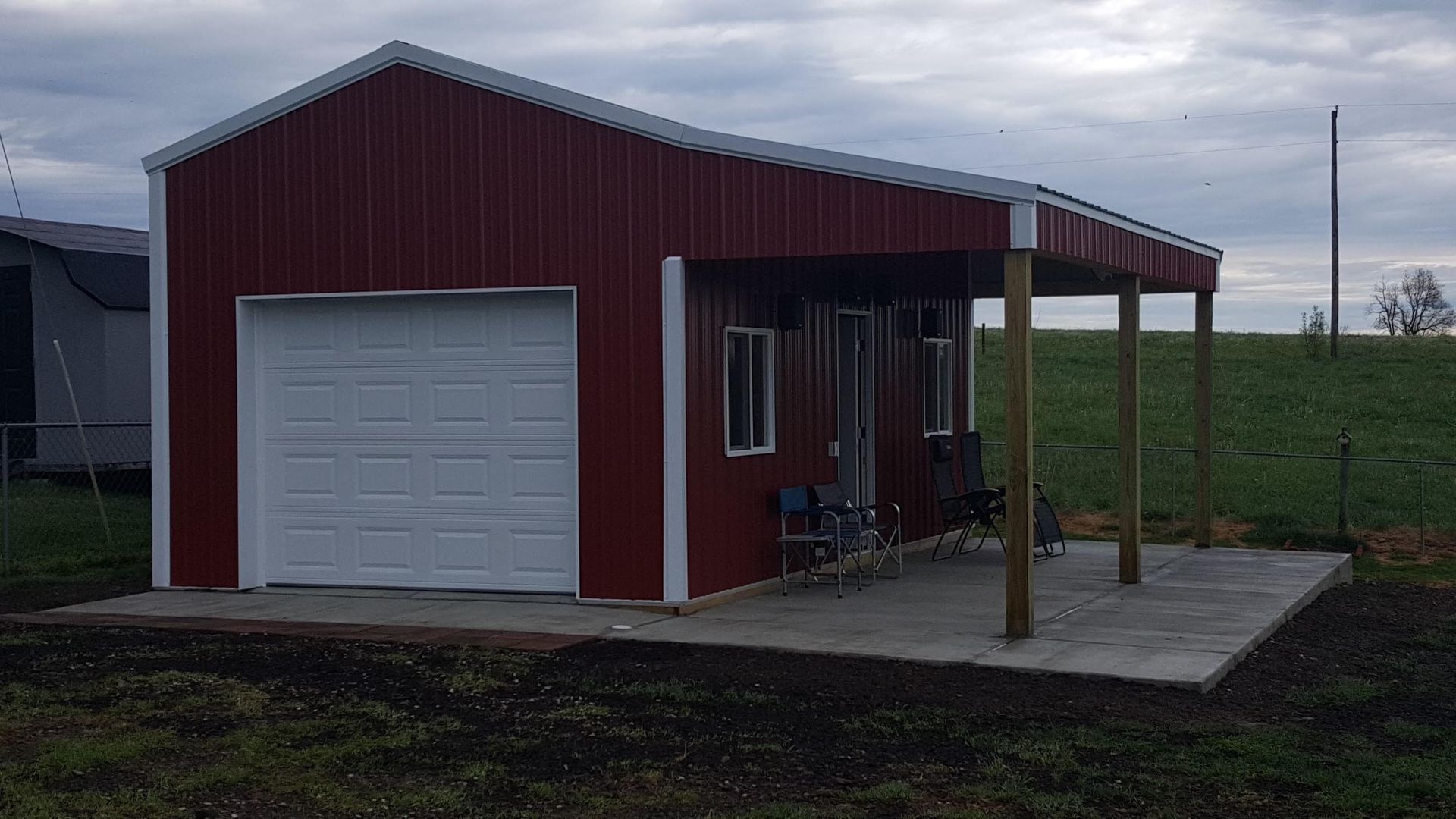 A red metal building with a garage door and an attached open porch on a concrete slab in a grassy field.