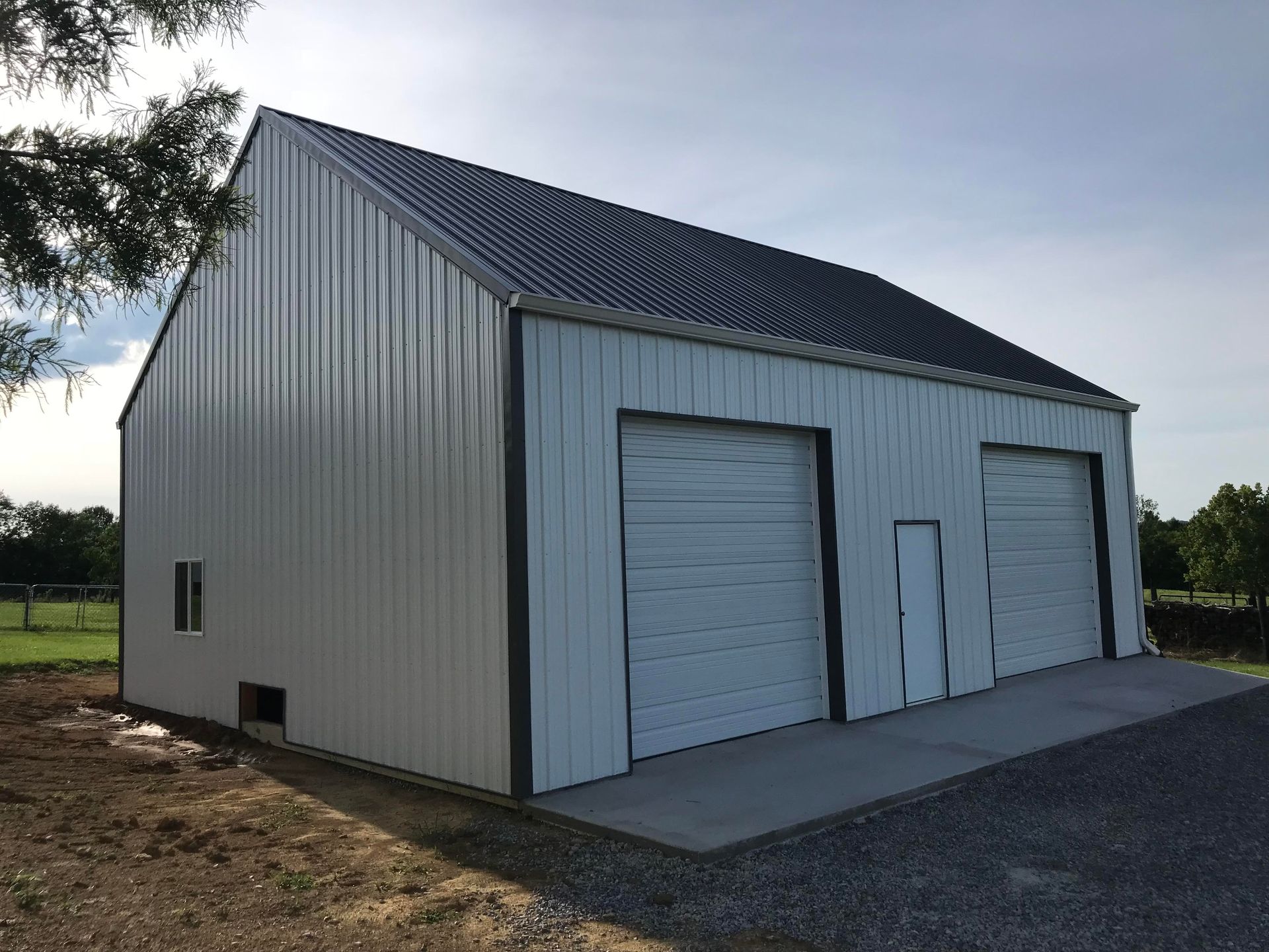 A white metal barn with a dark gray roof, two garage doors, and a side entry door, set in an open field.