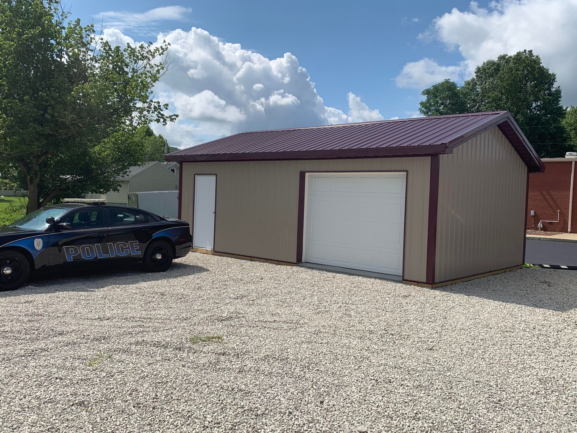 A tan metal garage with a dark roof and white doors parked next to a police cruiser on a gravel lot.