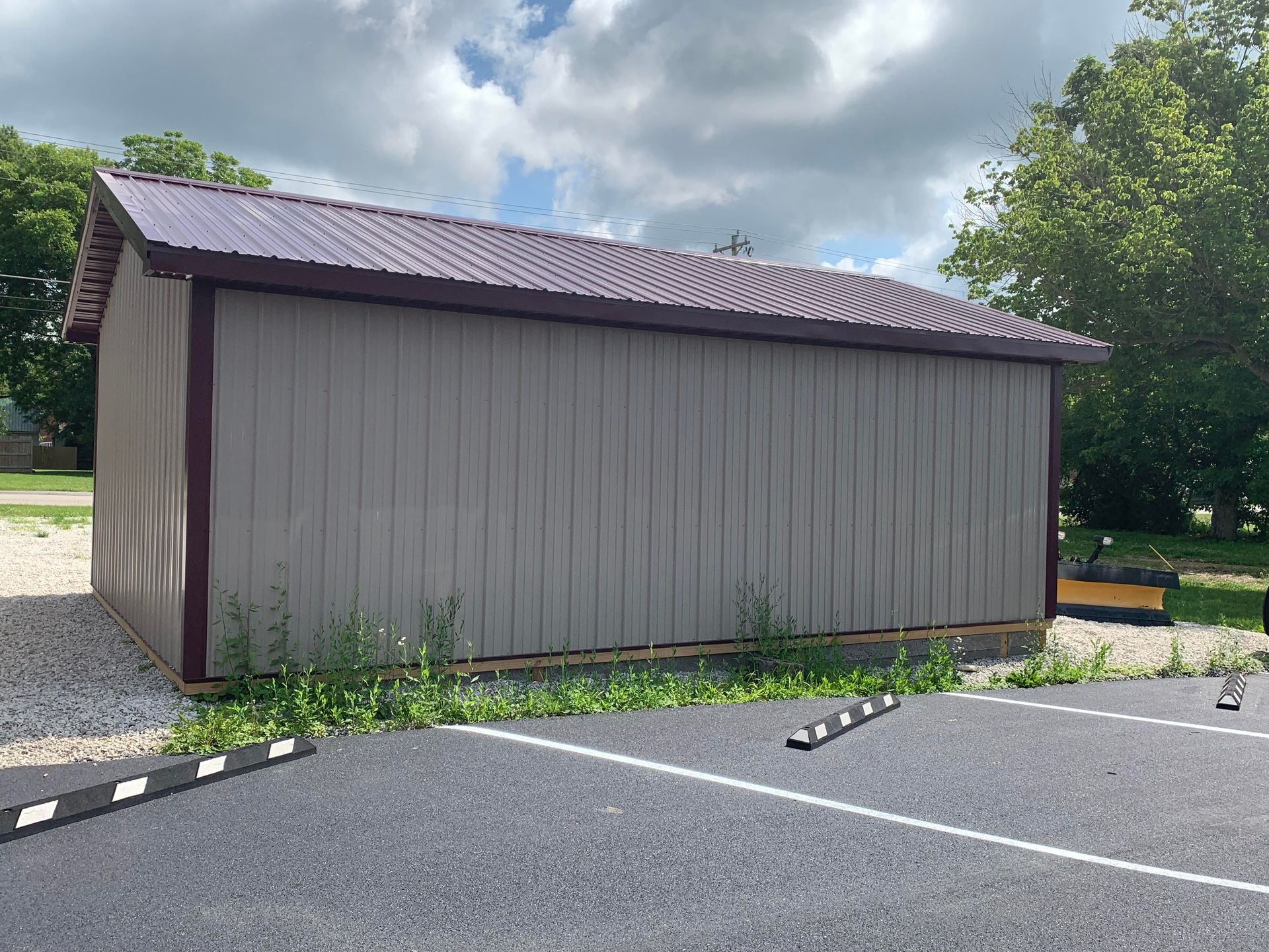 A tan metal storage shed with a dark roof sits on a gravel lot next to an asphalt parking area with white painted lines.