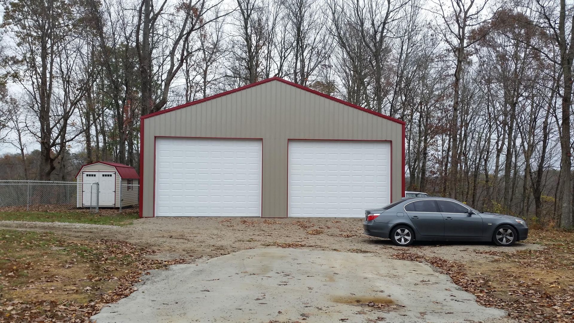 A large, beige metal garage with two white doors and a red roof, parked next to a small shed in a wooded area.