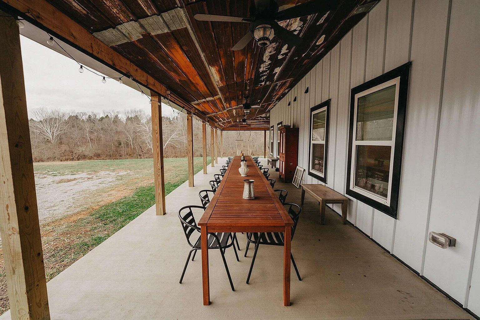 A long wooden dining table and chairs on a covered porch of a white farmhouse, looking out over an open, rural field.
