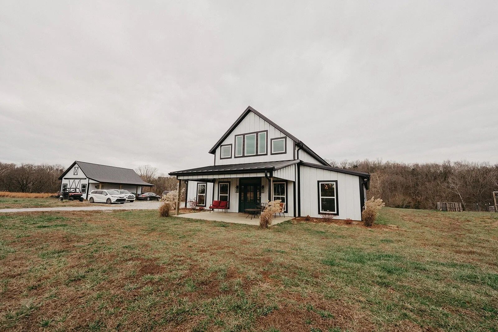 A white house with a dark metal roof and a wrap-around porch sits on a grassy field under a cloudy, overcast sky.