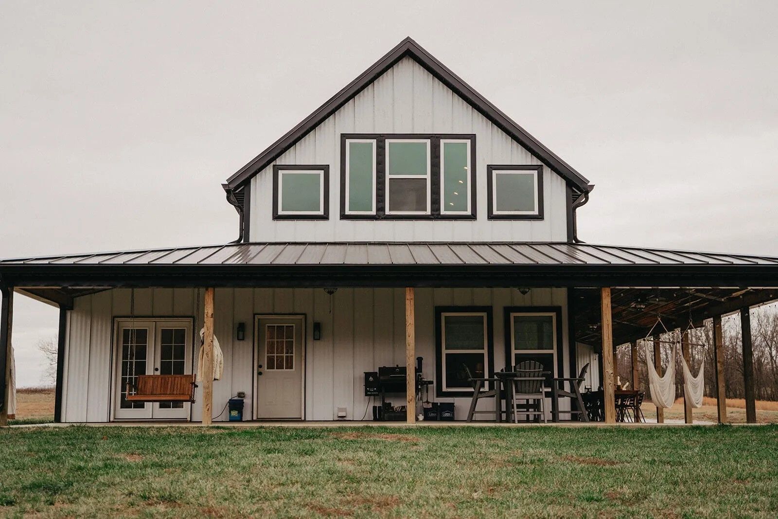 A white farmhouse with a black metal roof, front porch, and dark-trimmed windows set against a grassy field.