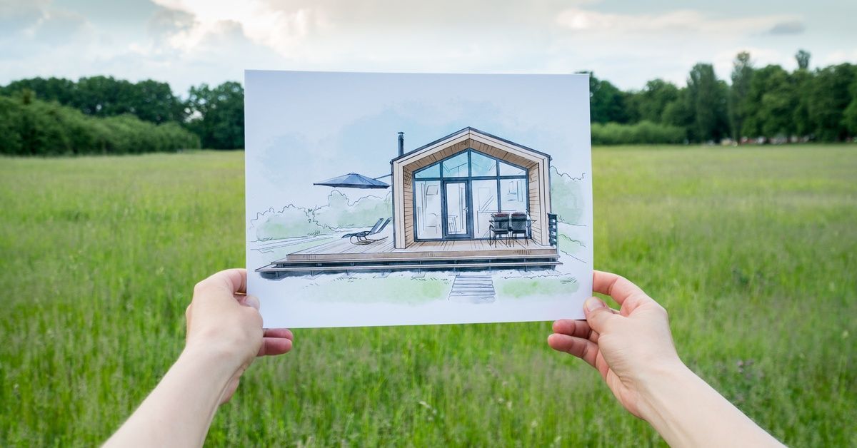 A person's outstretched hands hold a drawing of a pole barn house with a field, trees, and a partly cloudy sky in the background.