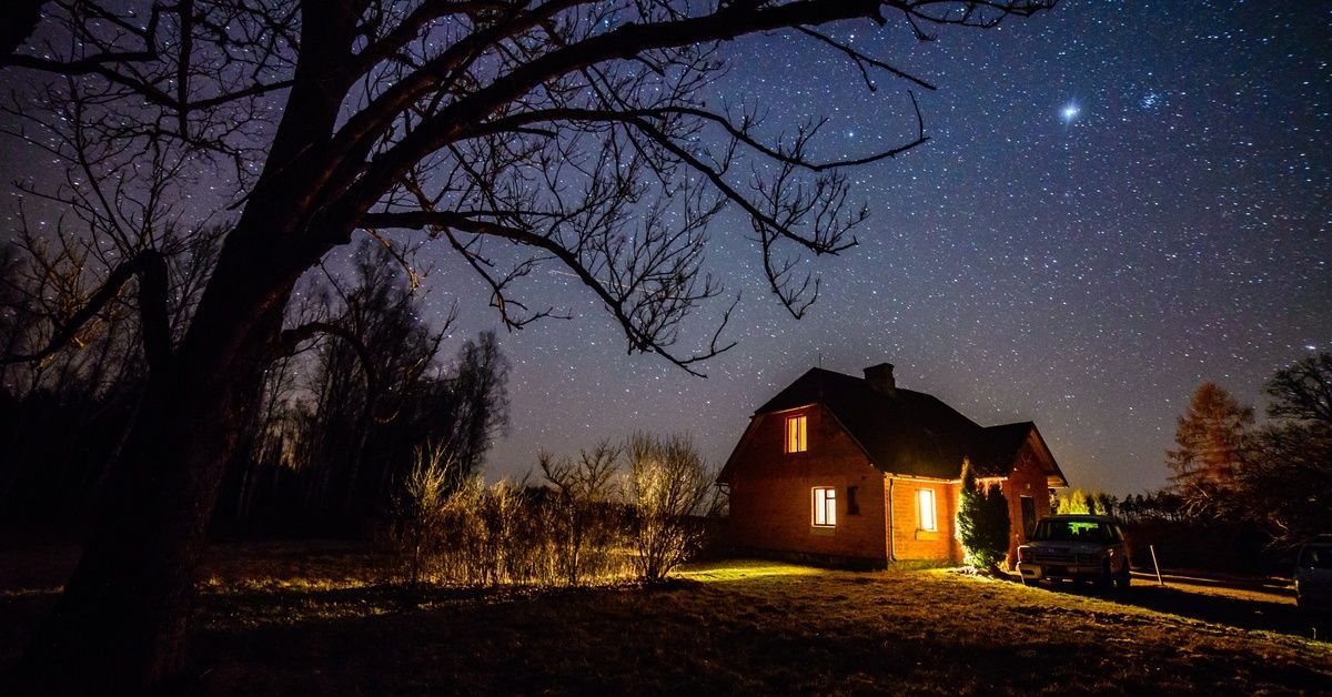 Stars shine brightly over a barn house with a car parked in front of it at night. Lights are on in the windows.