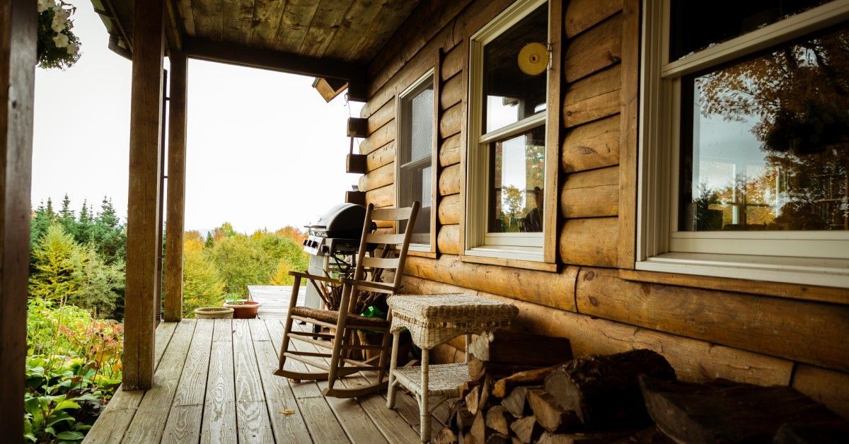 A wooden porch attached to a log cabin with several windows. A rocking chair, a grill, and a pile of wood sit on the porch.