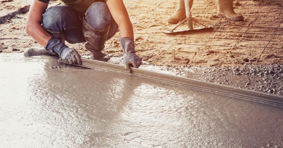 Construction workers smooth wet concrete on a foundation, with rebar visible in the background under