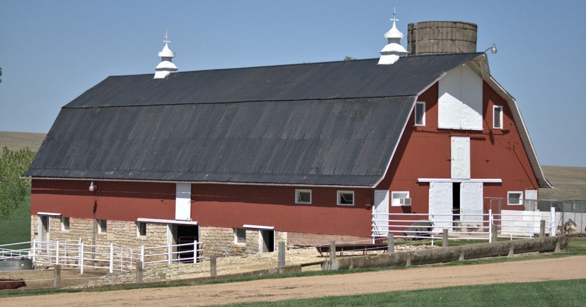 A red barn with a black metal roof sits on open farmland, featuring white trim, cupolas, fencing, and stone walls.