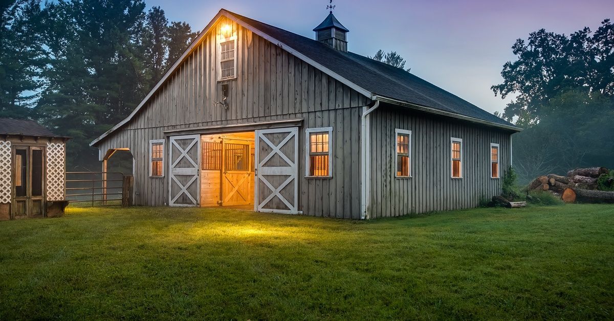 A large gray barn in a lush field at dusk. An exterior light is on outside of the barn, and the windows and door show light inside.