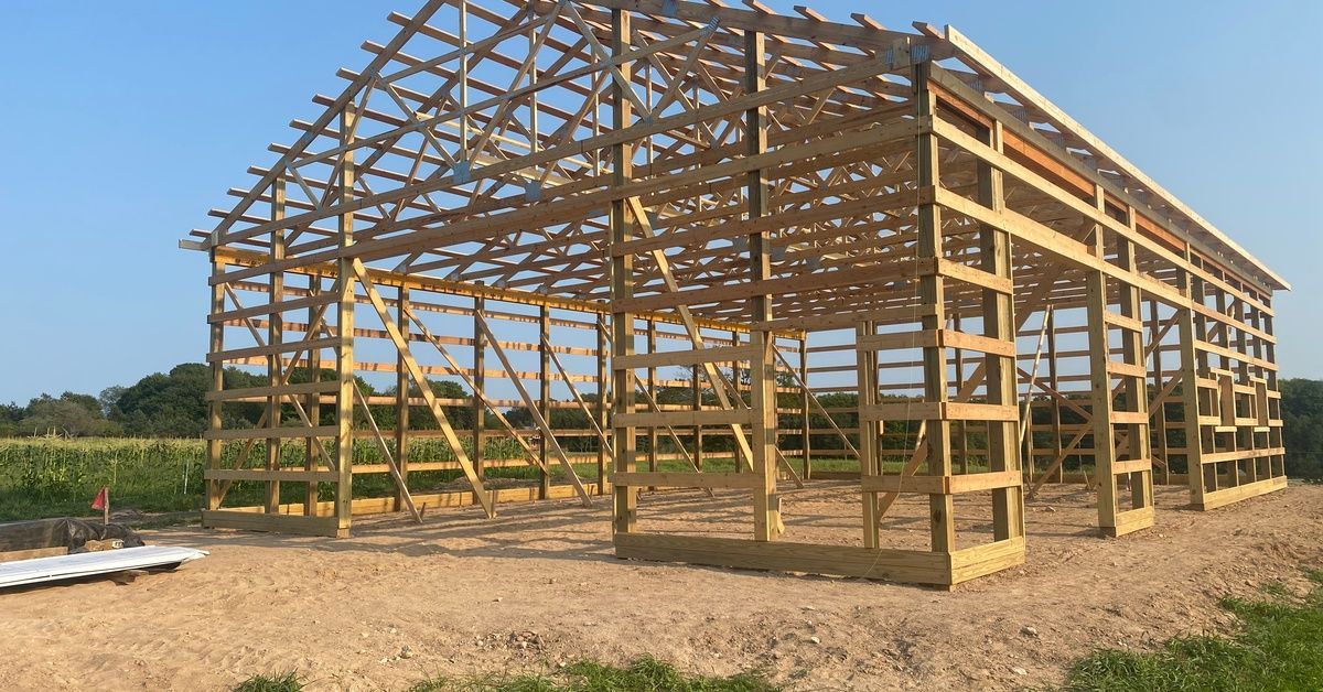 The bare, wooden framing of a barn house sitting on a patch of dry, sandy dirt, surrounded by grass.