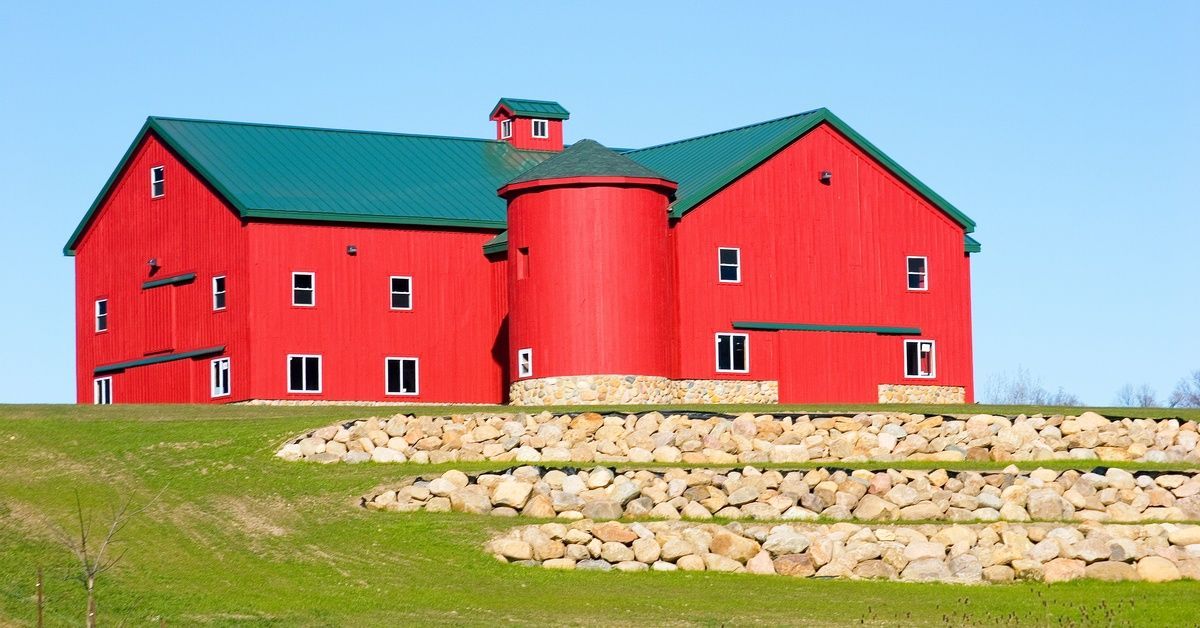A massive red barn with a green roof on a grassy, rural landscape. It is on top of a hill with lines of rocks nearby.