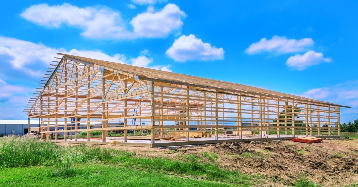 The wooden frame of a large pole barn with a pitched roof. The foundation of the barn is a concrete slab.