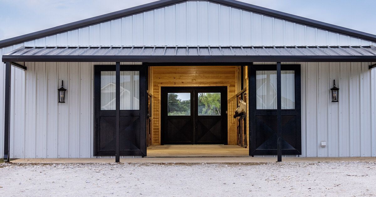 The exterior of a white metal barn with black accents and open double doors. There are horses inside the barn.