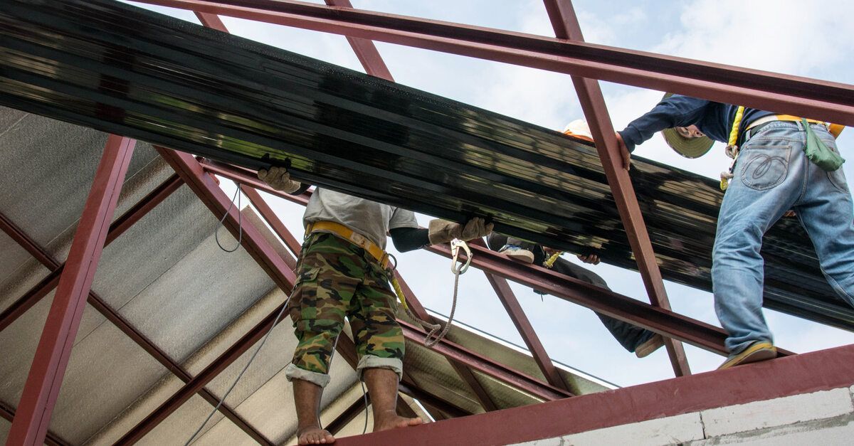 Three construction workers standing on a ledge and attaching a large metal sheet to a peaked barn roof.