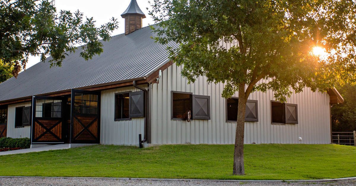 The exterior of a white metal barn with multiple windows. There is a turret on the roof and a horse looking out a window.