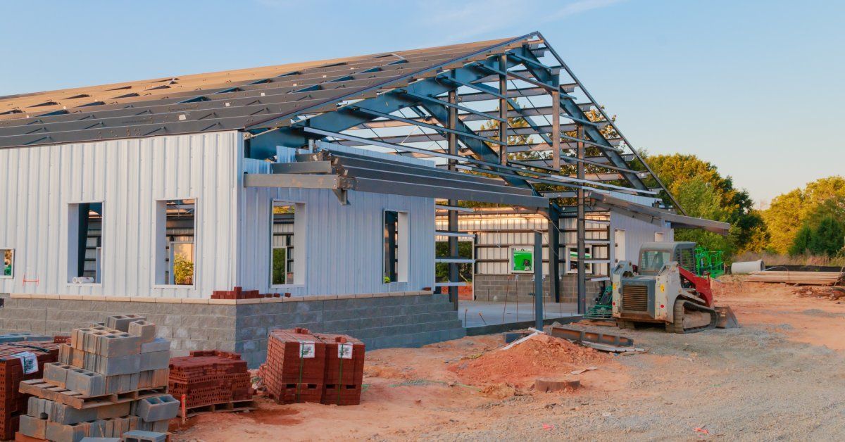 A large building being assembled with concrete blocks and white metal siding. There is a small excavator in front of the building.