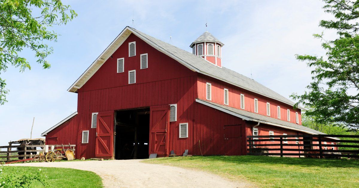 The exterior of a large barn with red siding, a peaked roof with a cupola, and several white windows.