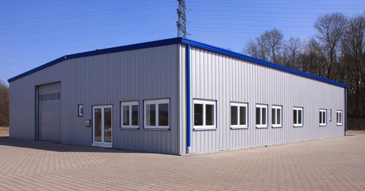 A gray metal building with blue trim and several windows surrounded by a brick driveway and mature trees.
