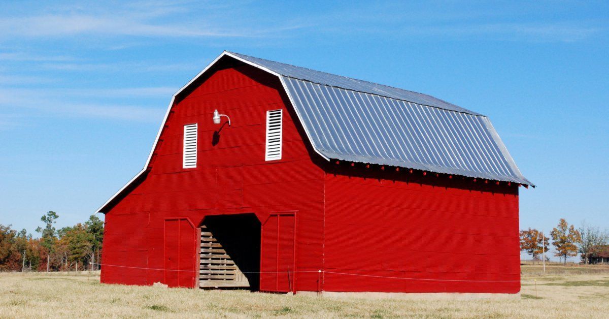 The exterior of a bright red barn with a gray metal roof. The front of the barn has two white windows and a large open door.